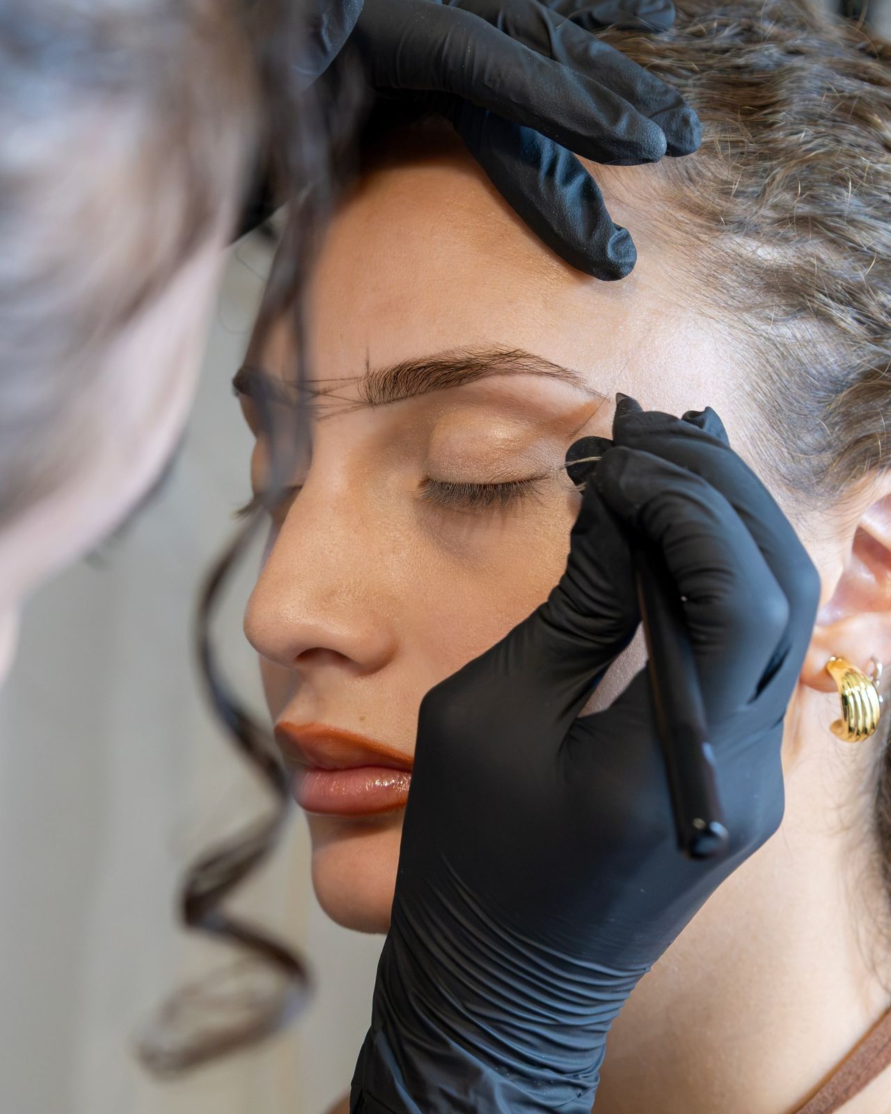 Con il guanto nero, Morgana disegna con la matita la forma delle sopracciglia sul viso della cliente, a occhi chiusi